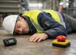Industrial worker in a hard hat and high-visibility vest lying on the factory floor, with a red emergency stop button and a handheld device nearby.