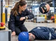 Woman kneeling beside a man lying on the factory floor, checking her watch during a medical emergency drill in a warehouse.